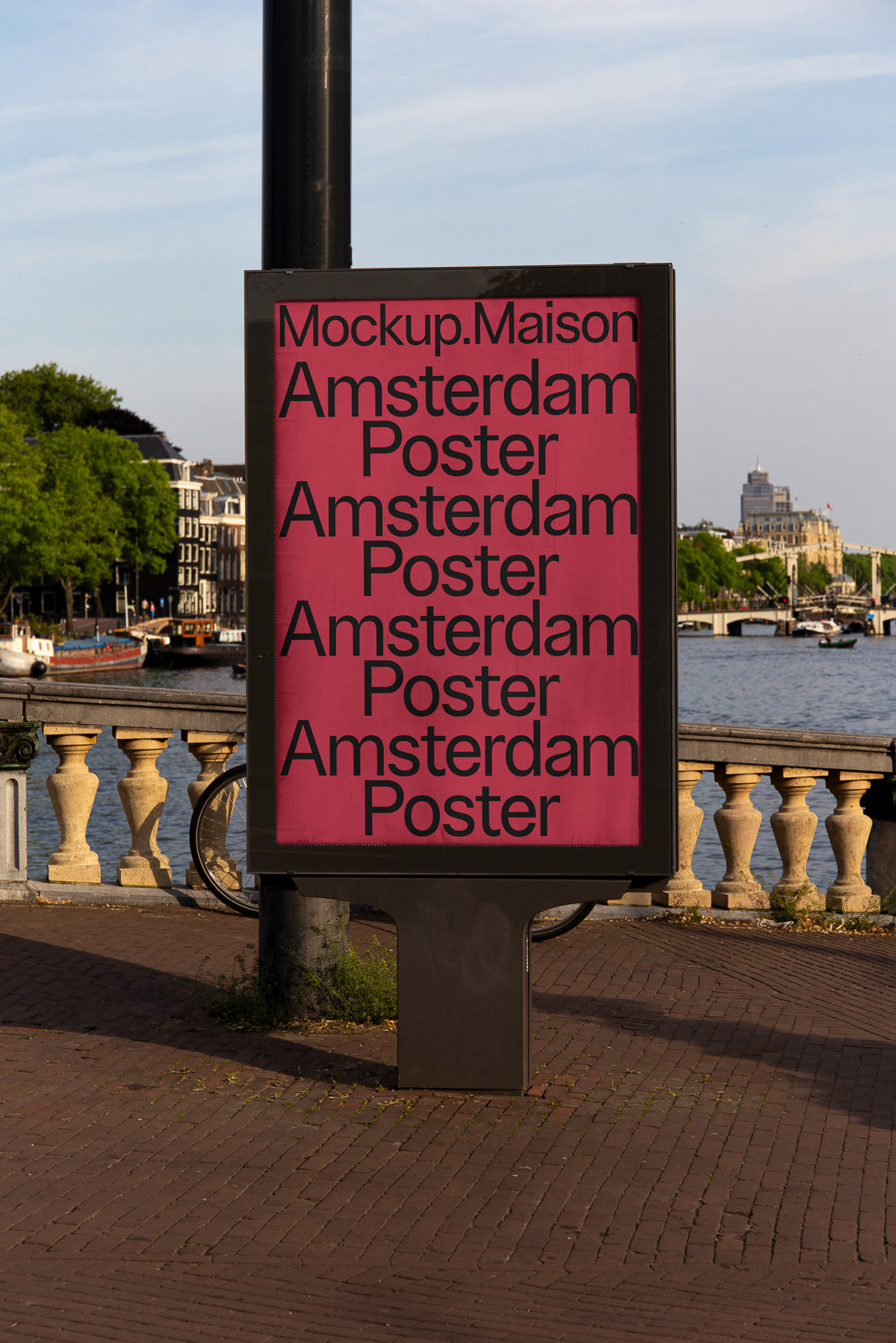 PSD mockup of a large outdoor poster display on a city street corner near a canal, featuring repetitive "Amsterdam Poster" text on a pink background.