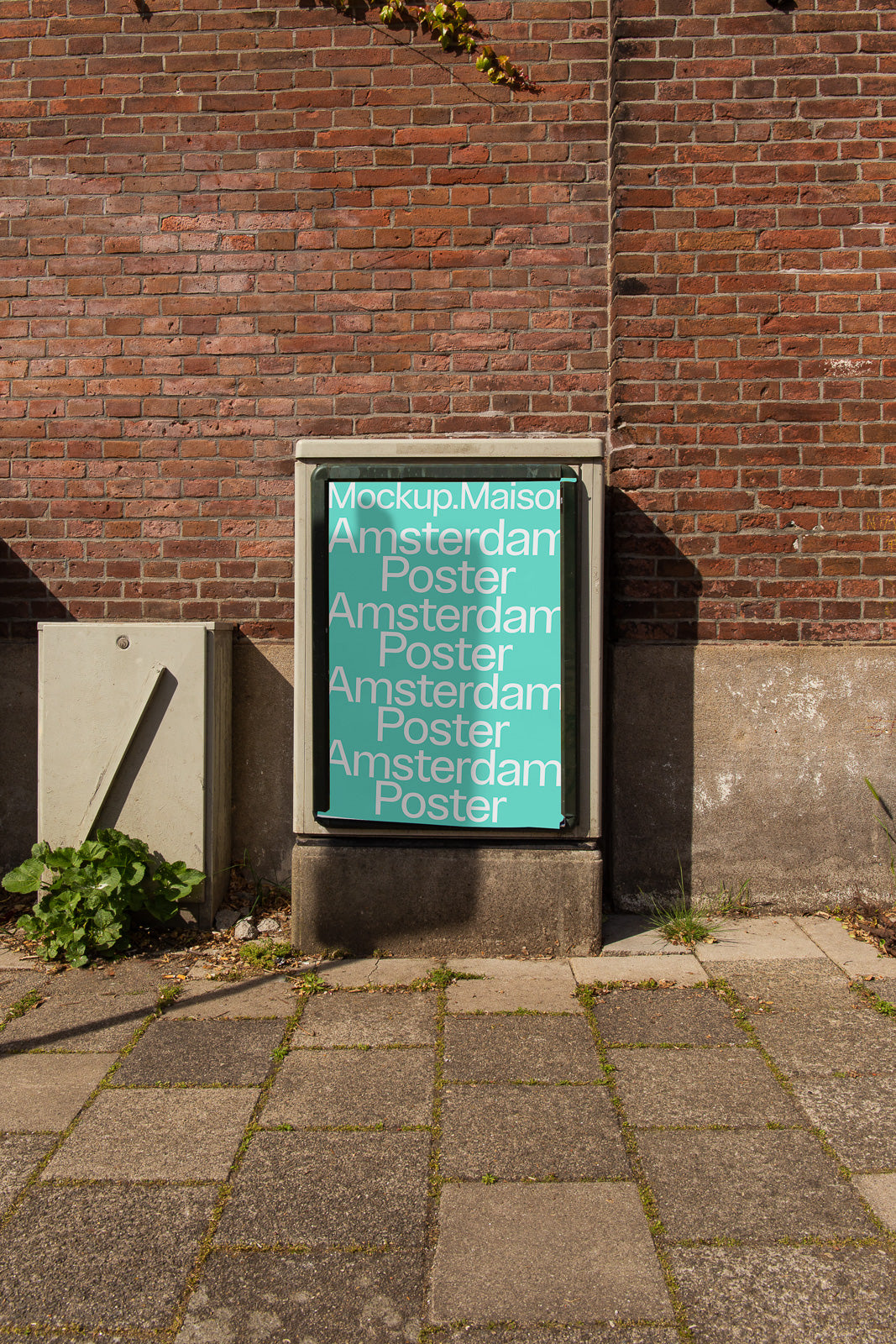 PSD mockup of a vertical poster showcasing text "Mockup.Maison Amsterdam Poster" placed against a brick wall outdoors.