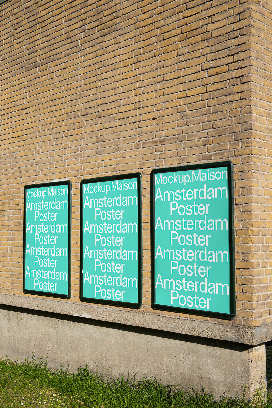 PSD mockup of three vertical posters on a brick wall, each displaying "Mockup.Maison Amsterdam Poster." The posters are framed and set above green grass.