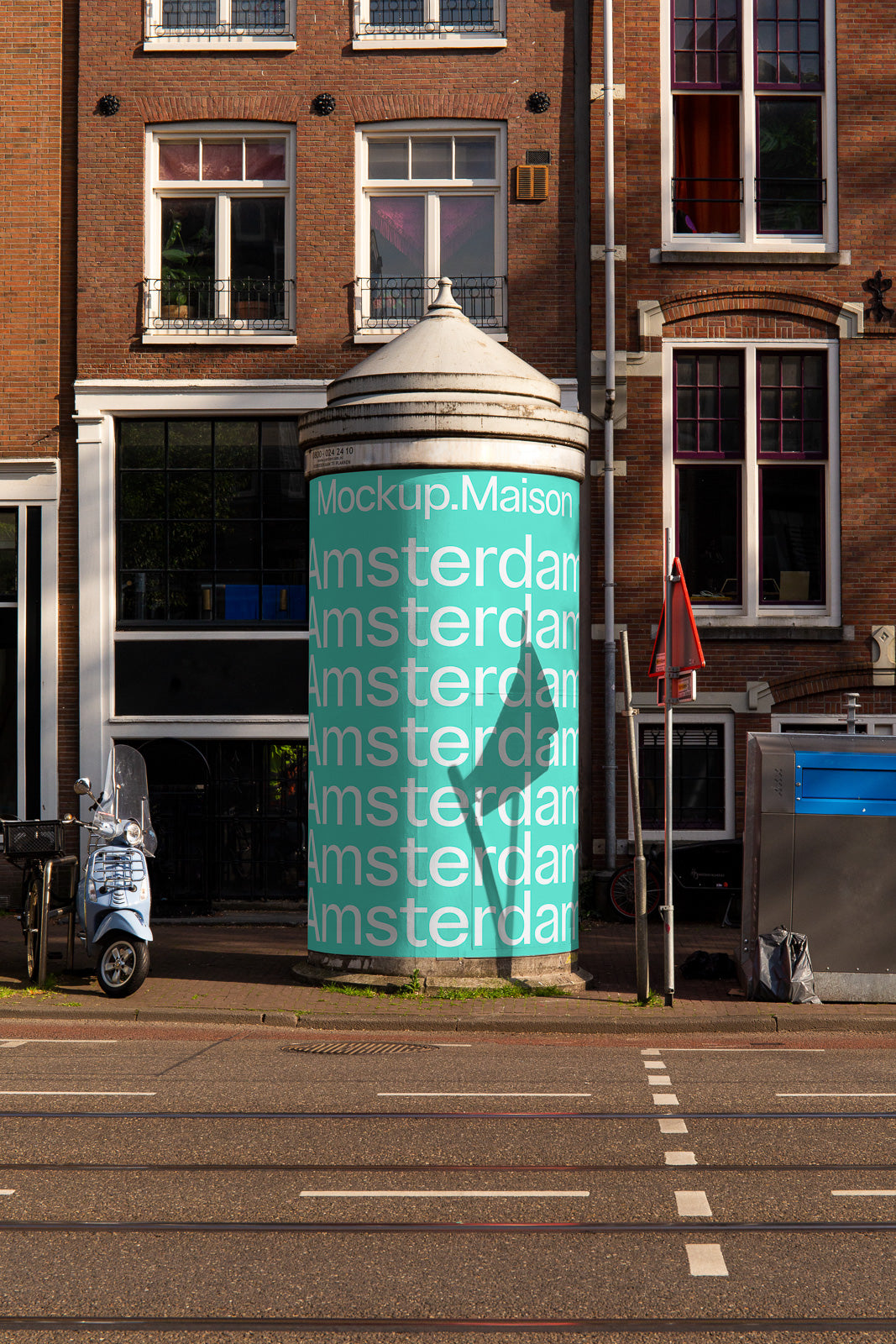 PSD mockup of a large cylindrical structure with repeating "Amsterdam" text and "Mockup.Maison" on a teal background, set against a brick building.