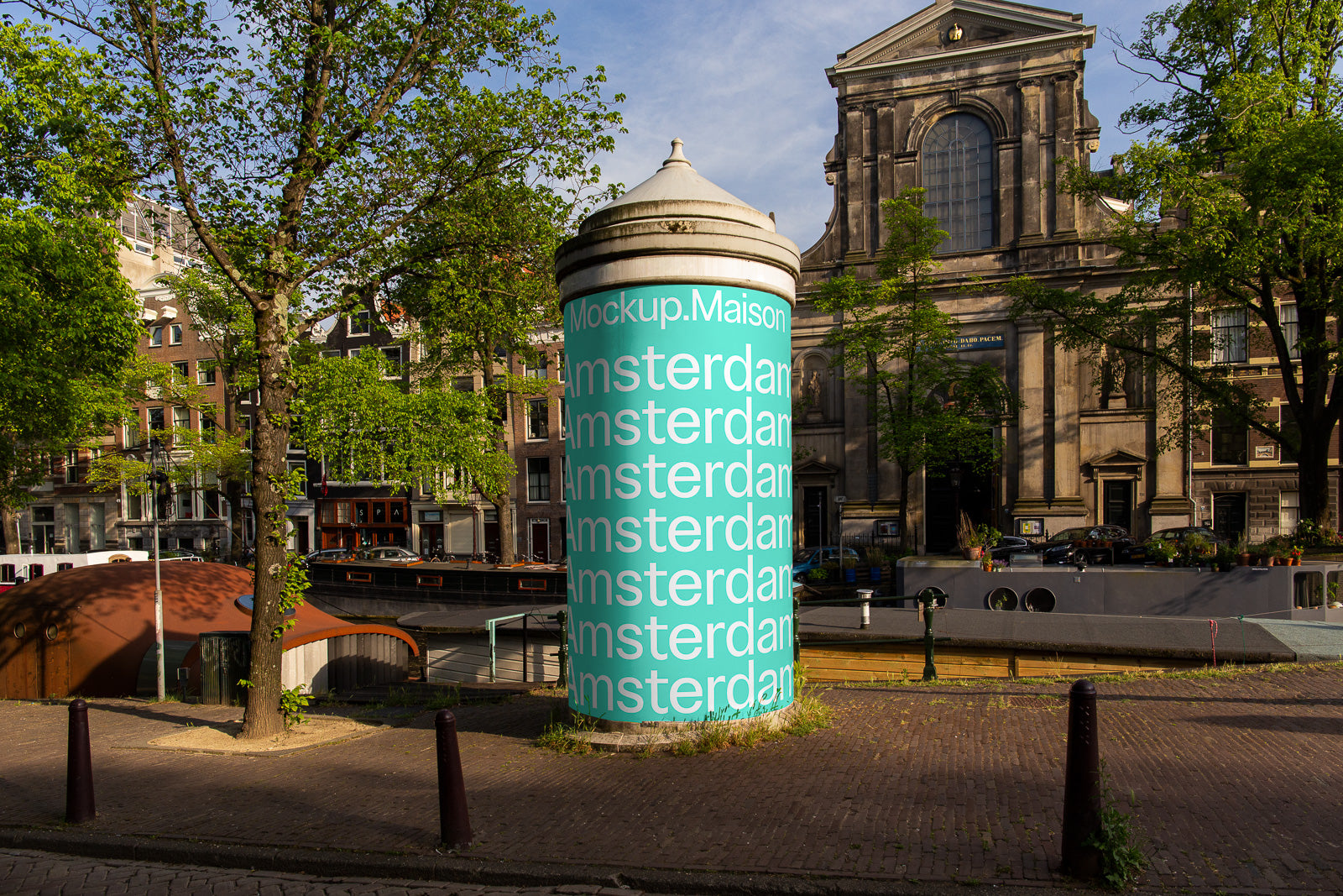 PSD mockup of a cylindrical advertisement pillar displaying the word "Amsterdam" repeatedly, situated in a city street with trees, buildings, and a canal.