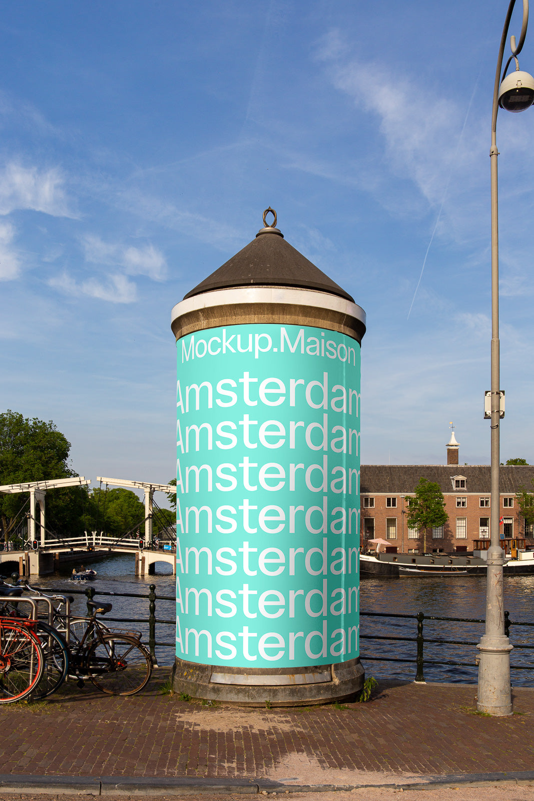 PSD mockup of a cylindrical column near a canal, wrapped with turquoise "Amsterdam" text, under a clear blue sky with bridge and trees in view.