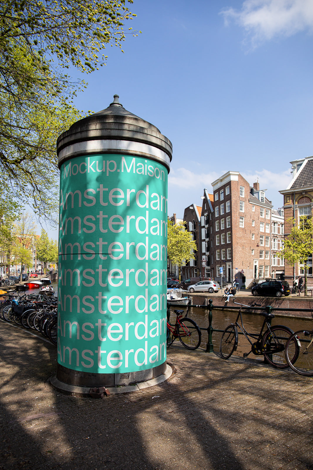 PSD mockup of a cylindrical advertising column in an urban setting, showcasing a bold, turquoise design with repeated text "Amsterdam" against a canal backdrop.