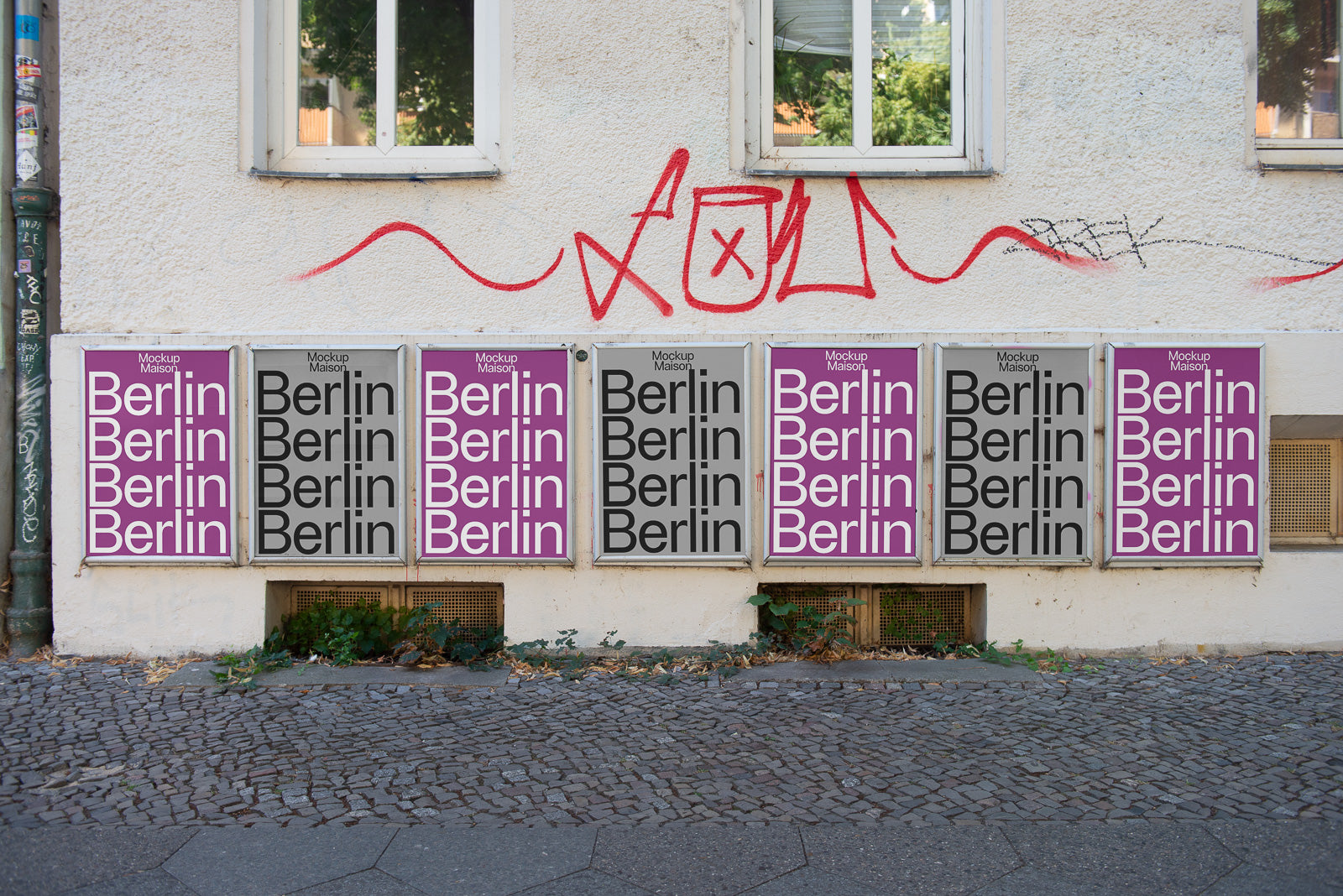 PSD mockup of posters on an urban wall, featuring repeated "Berlin" text in variations of purple and gray, with graffiti above and cobblestone sidewalk below.