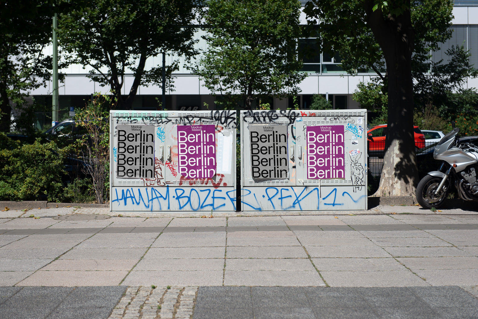 PSD mockup of street utility box covered with Berlin posters and graffiti, located on a sidewalk with trees and a parked motorcycle nearby.