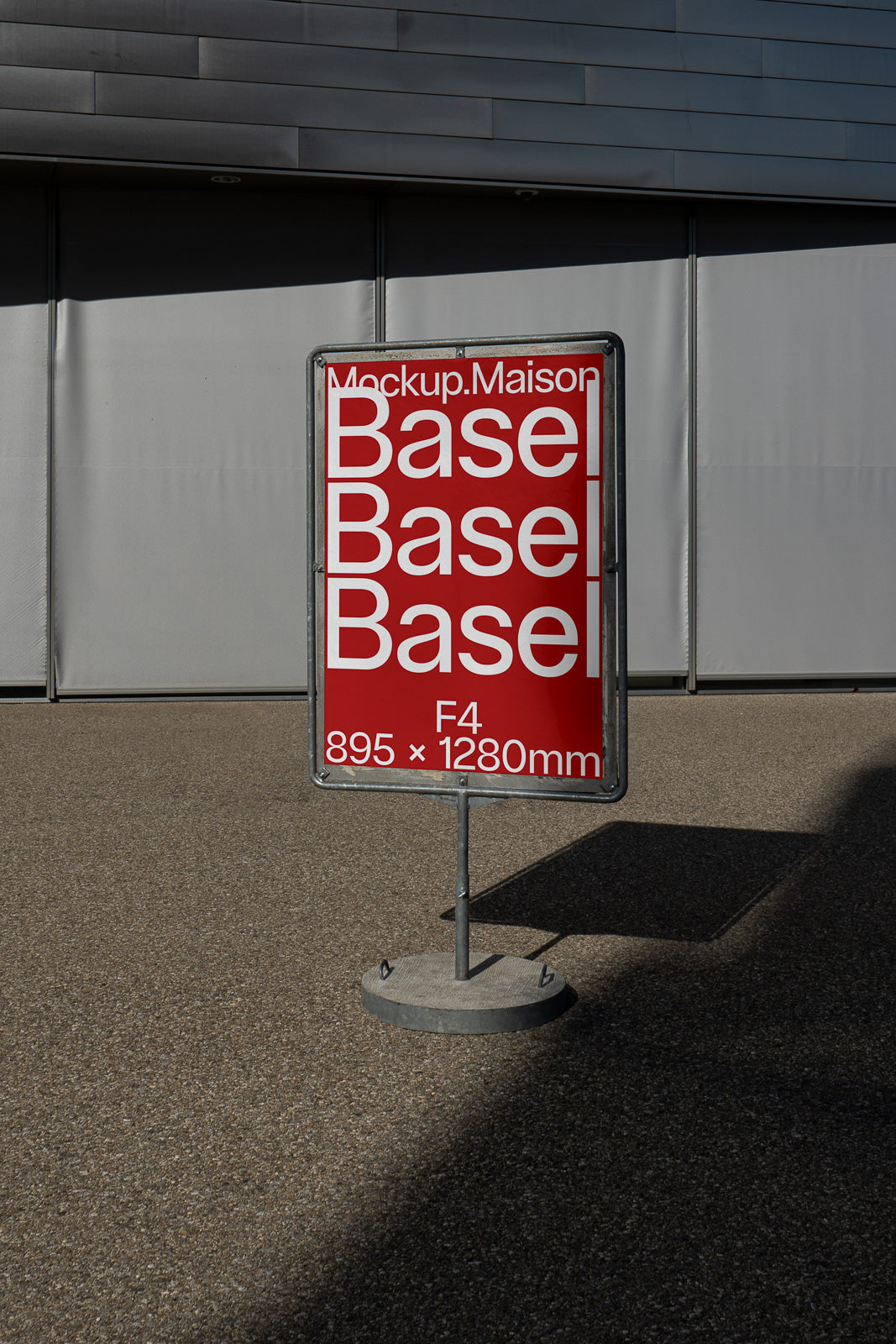 PSD mockup of a freestanding metal sign with a red poster displaying the words "Base Basel" in white text, positioned outside a modern building.
