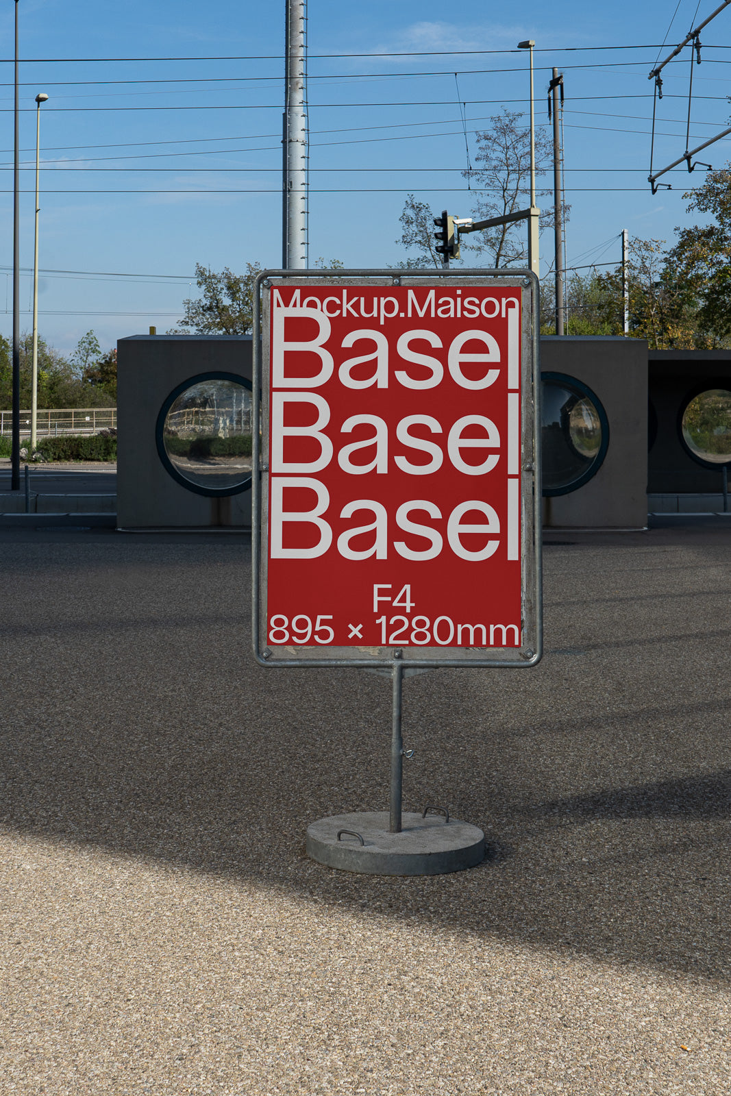 PSD mockup of a street sign displaying text "Mockup.Maison Basel Basel Basel F4 895 × 1280mm" in front of an urban backdrop with poles and circular windows.