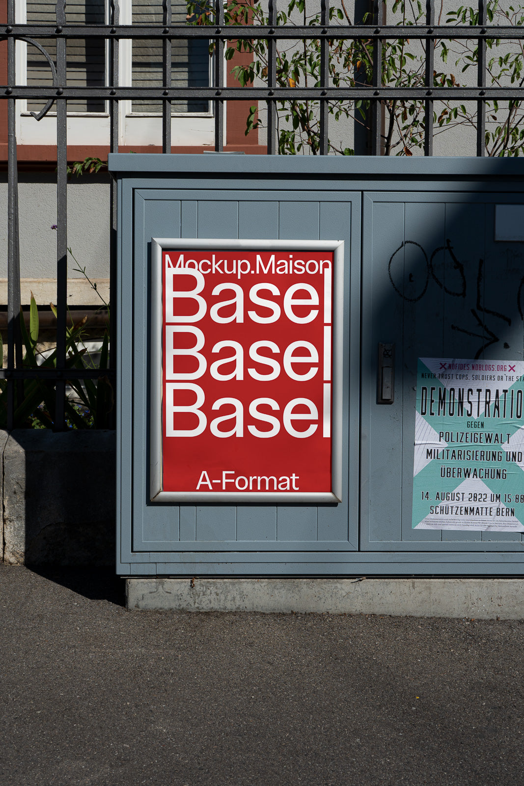 PSD mockup of a poster labeled "Mockup.Maison Basel" in white text on a red background, displayed on an outdoor cabinet in an urban setting.