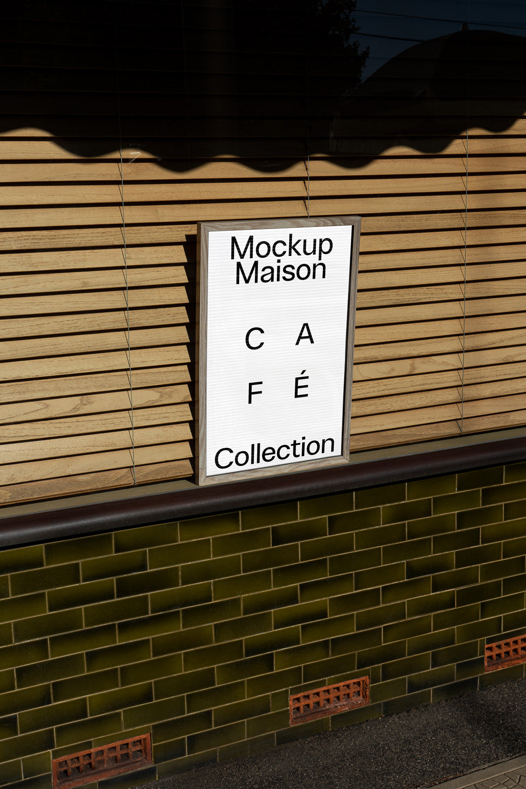 PSD mockup of a framed poster leaning against a wall with wooden slats and green tiles, displaying the text "Mockup Maison Café Collection."