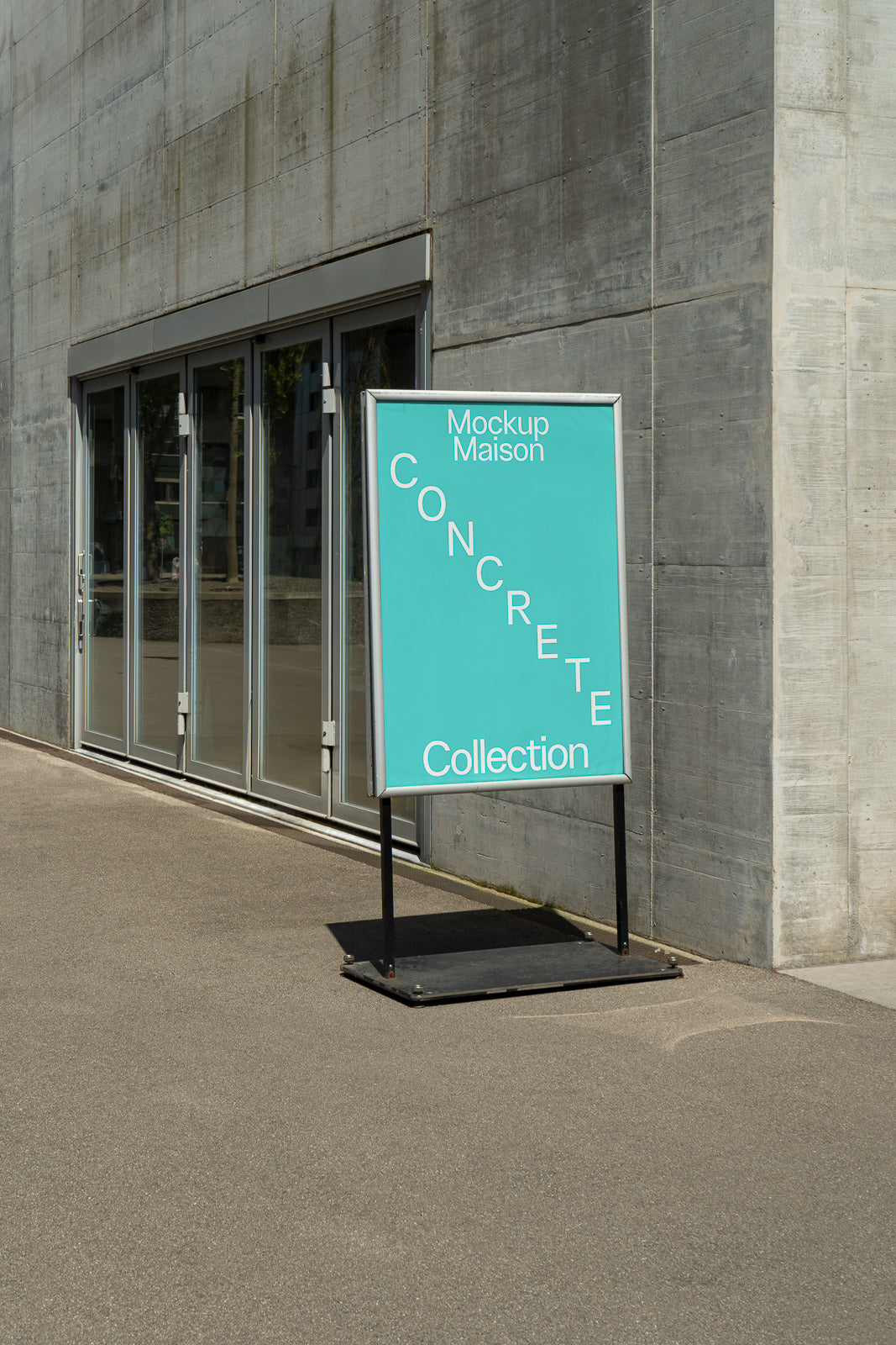 PSD mockup of a sidewalk sign with "Mockup Maison Concrete Collection" text, positioned next to a modern building's glass doors on a sunny day.