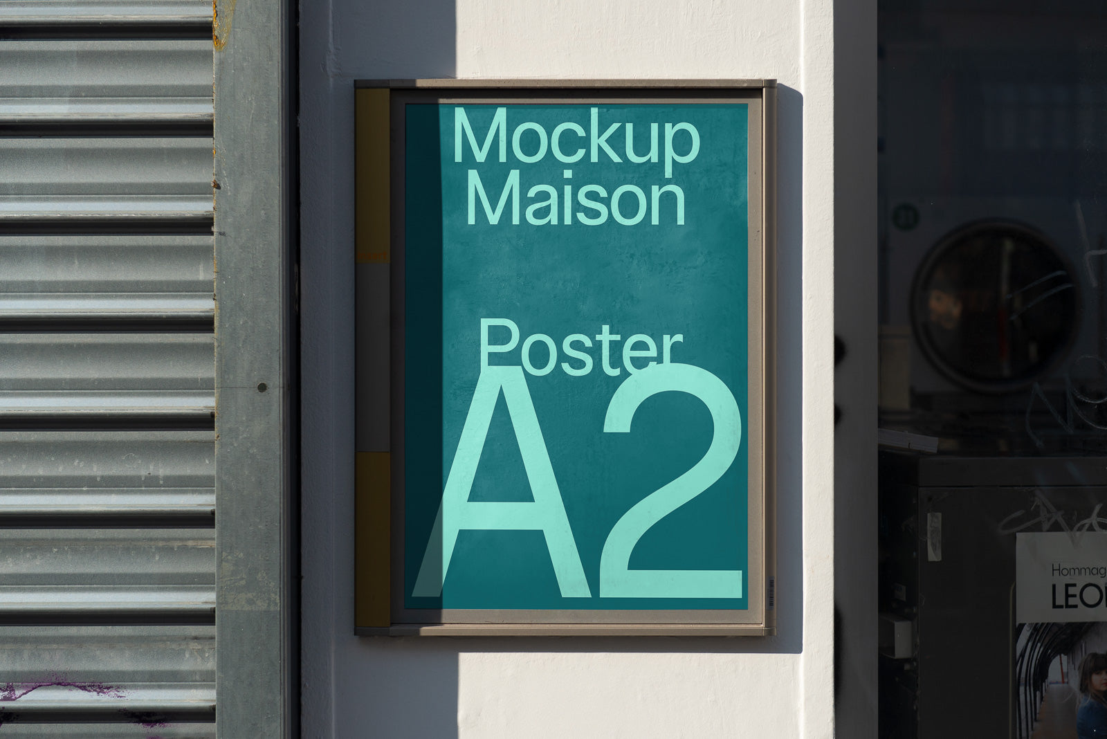 PSD mockup of an A2 poster displayed in a wooden frame on an exterior wall next to a metal shutter, with sunlight casting shadows.