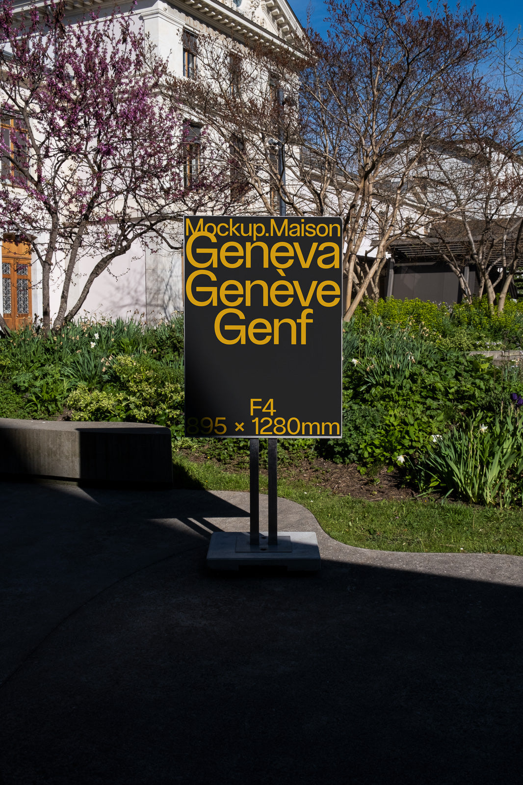 PSD mockup of an outdoor standing sign displaying text in black and yellow, situated in a garden setting with trees and a building in the background.