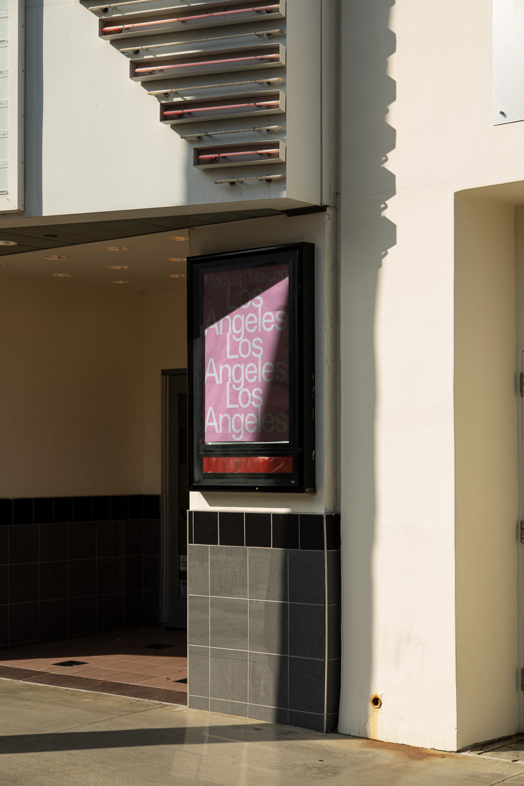 PSD mockup of a vertical poster frame mounted on a building's exterior wall, displaying layered text "Los Angeles" in light-colored fonts against a pink background.