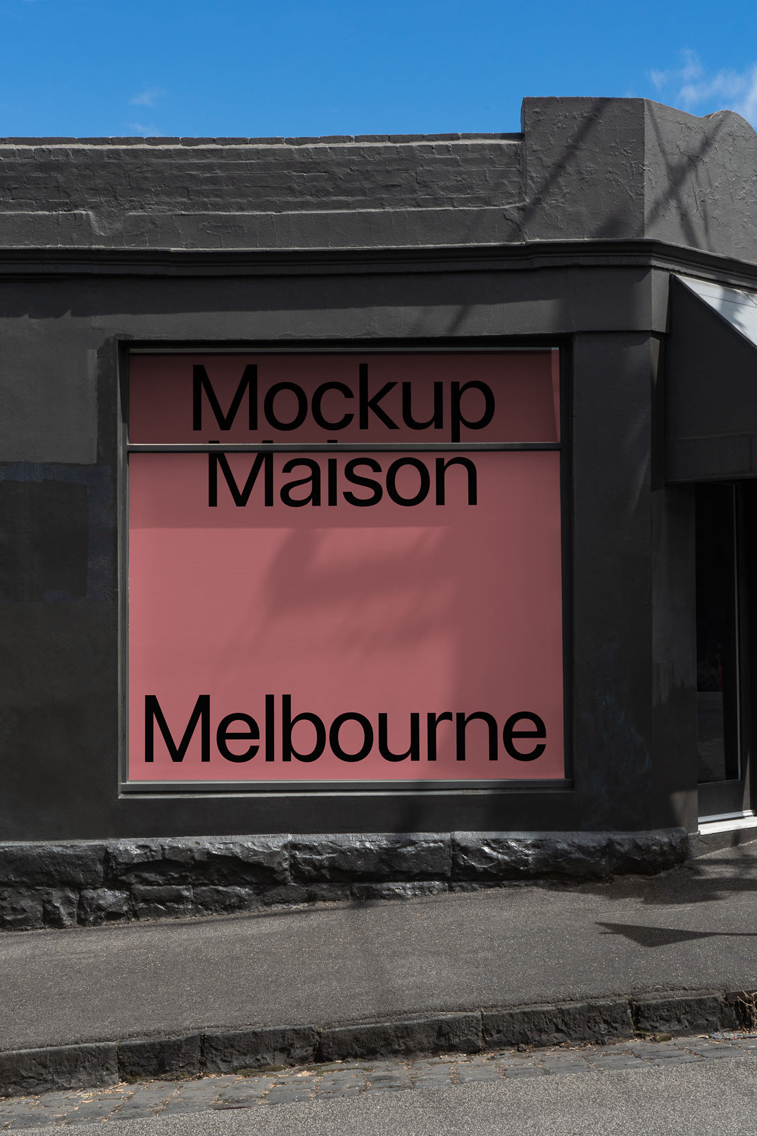 PSD mockup of a shopfront window with a pink background displaying large black text "Mockup Maison Melbourne" against a dark gray building facade.