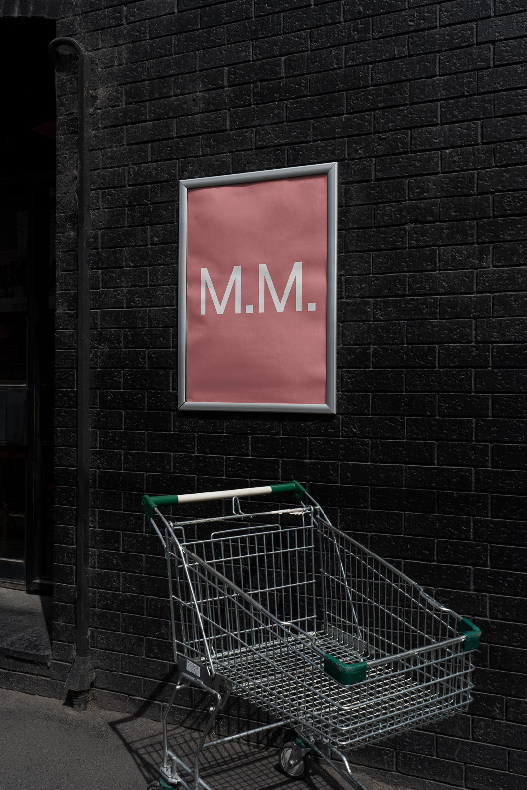 PSD mockup of a framed pink poster with "M.M." text on a black brick wall next to a metal shopping cart.