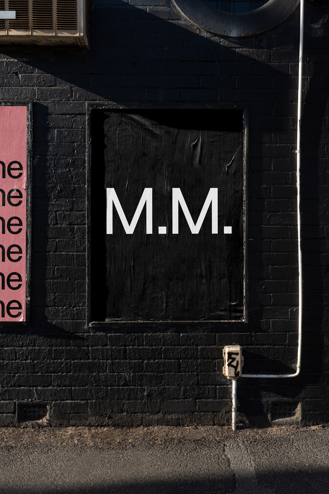 PSD mockup of a black brick wall featuring a white "M.M." centered on a black poster, adjacent to a pink poster with repeated text.