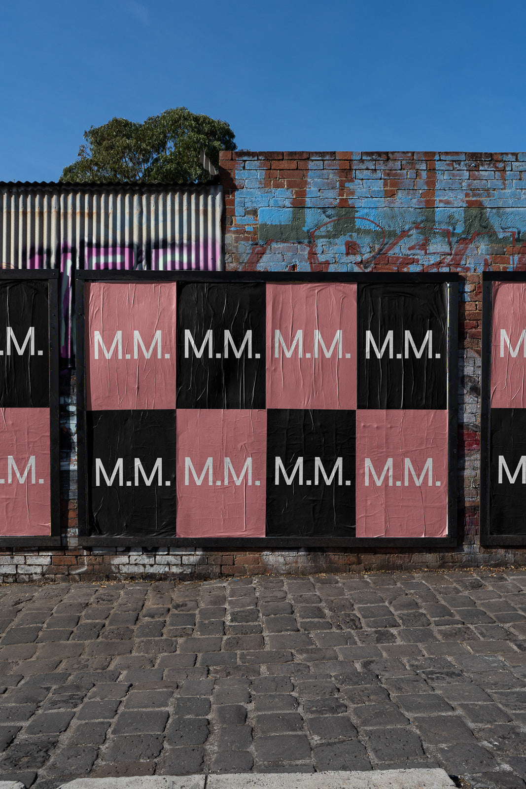 PSD mockup of street posters on a brick wall with alternating black and pink backgrounds featuring "M.M." in white text.