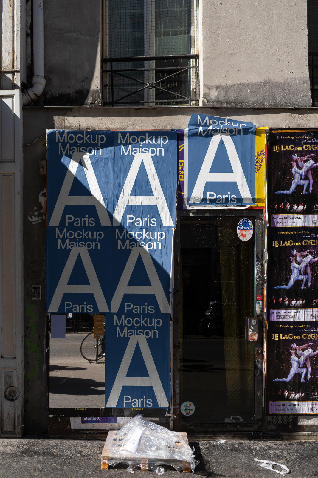 PSD mockup of a storefront wall covered with blue posters labeled "Mockup Maison A Paris" and a stack of wrapped pallets on the ground.