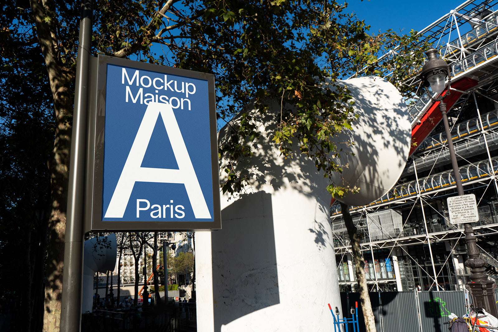 PSD mockup of an outdoor sign displaying blue and white text reading "Mockup Maison A Paris," set against an urban background with trees and a building.