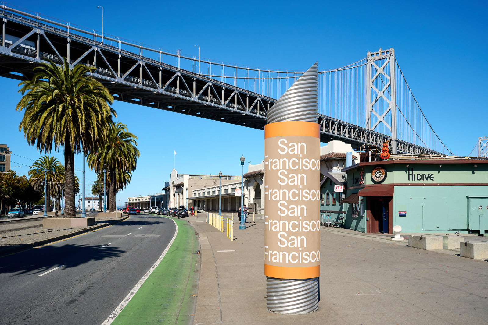 PSD mockup of San Francisco signage post under a bridge with clear blue sky, nearby palm trees, and surrounding buildings.