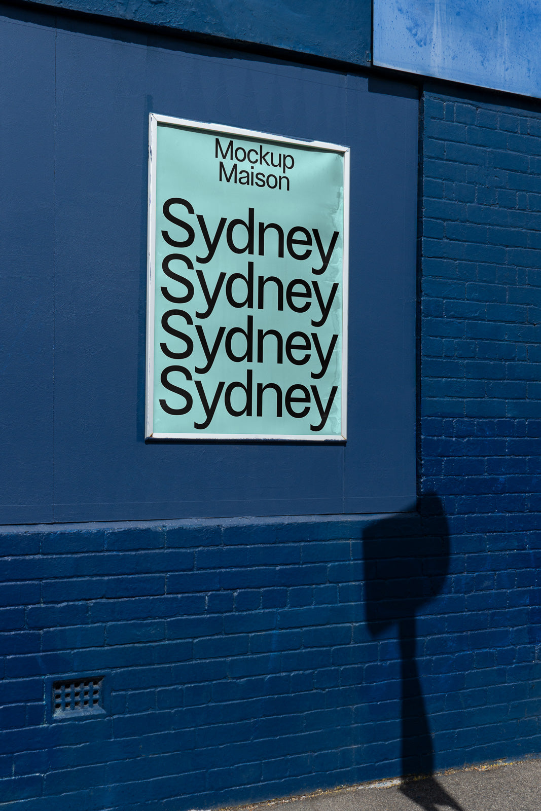 PSD mockup of a blue wall with a poster featuring repeated text "Sydney" in bold black letters. The poster is framed and contrasts against the deep blue bricks.
