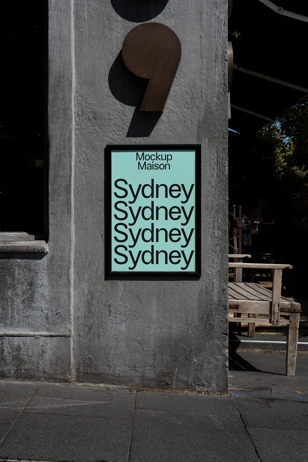 PSD mockup of a framed poster on an outdoor gray wall, featuring the repeated word "Sydney" in bold text with a shadow effect cast by a large comma sculpture above.