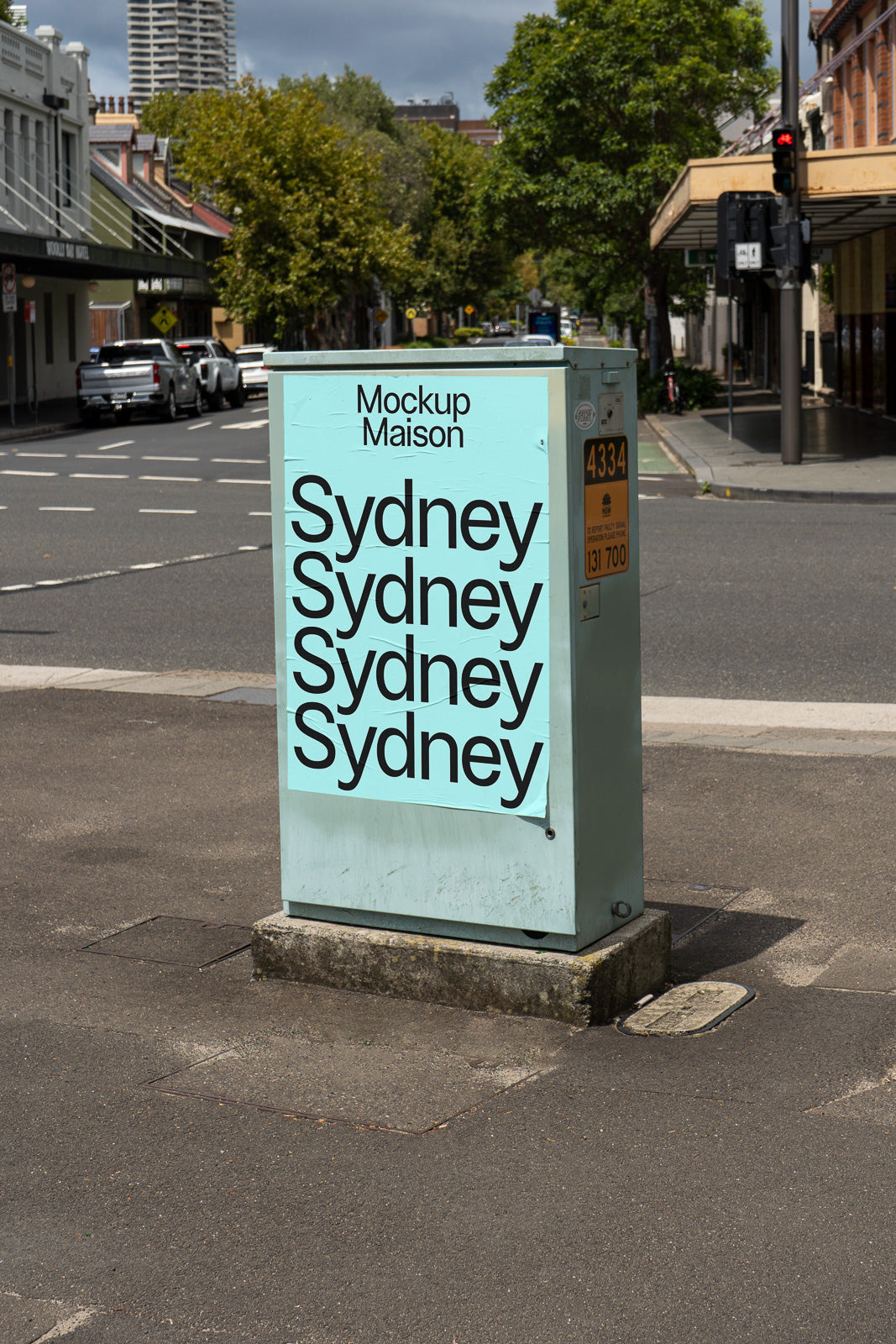 PSD mockup of a utility box on a street corner with a poster reading "Sydney" in large text, surrounded by urban buildings and trees.