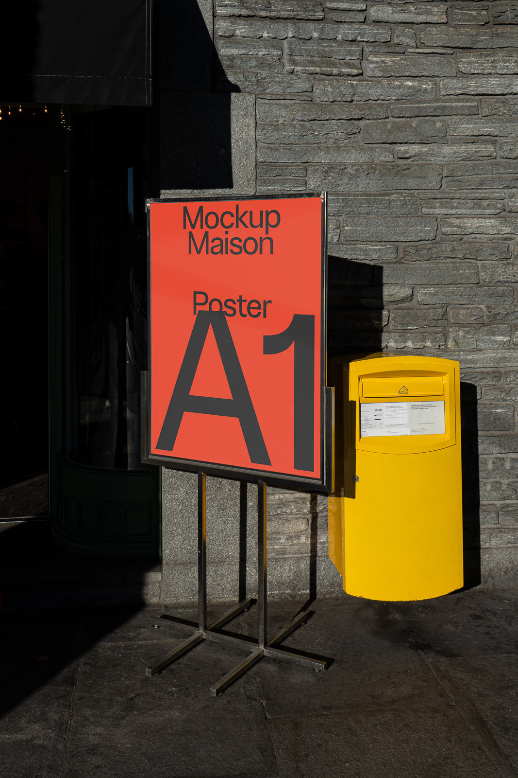 PSD mockup of a red A1 poster displayed on a stand next to a yellow mailbox against a gray stone wall.