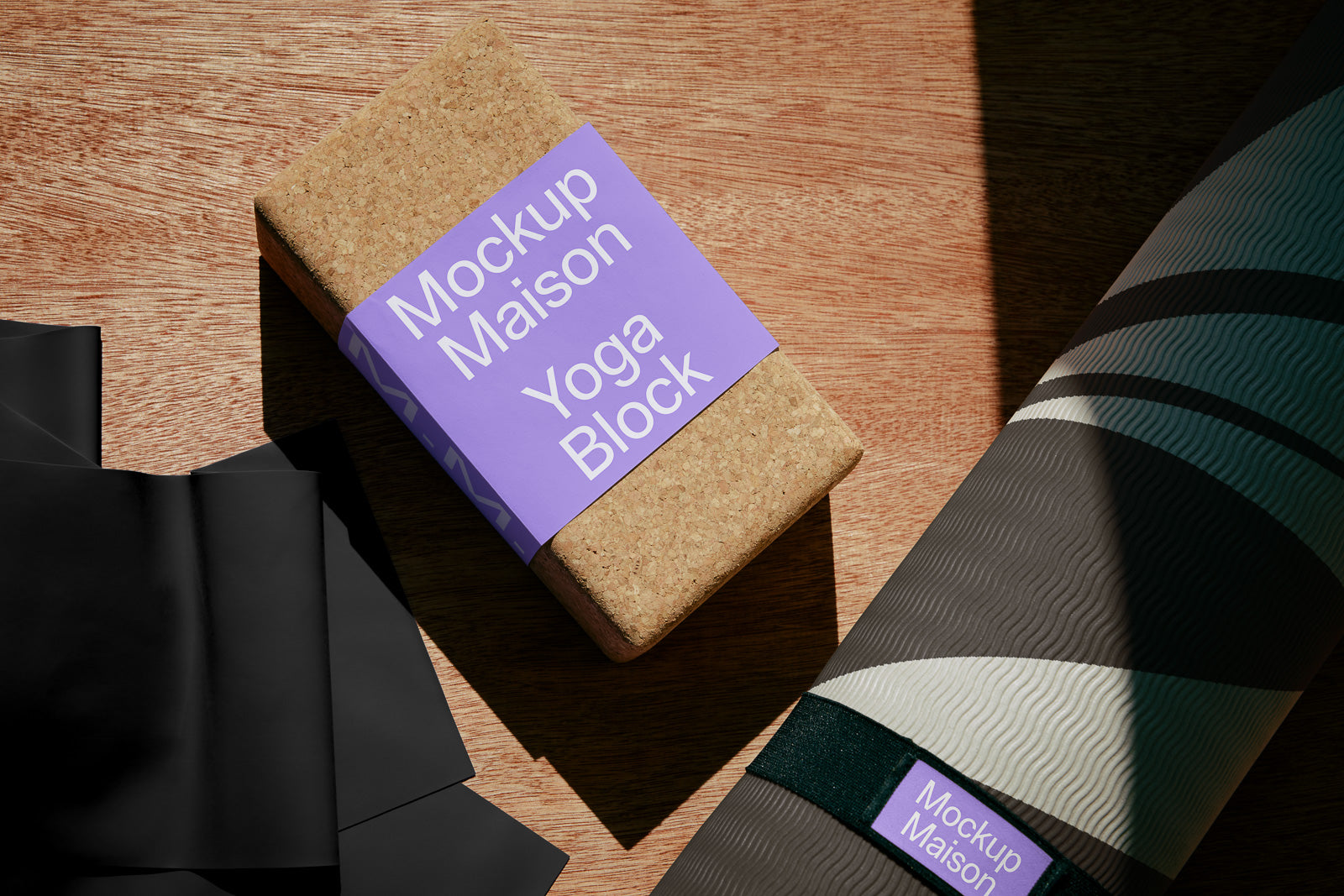 PSD mockup of a cork yoga block labeled "Mockup Maison Yoga Block" with a purple band, placed next to a yoga mat and black sheets on a wooden surface.