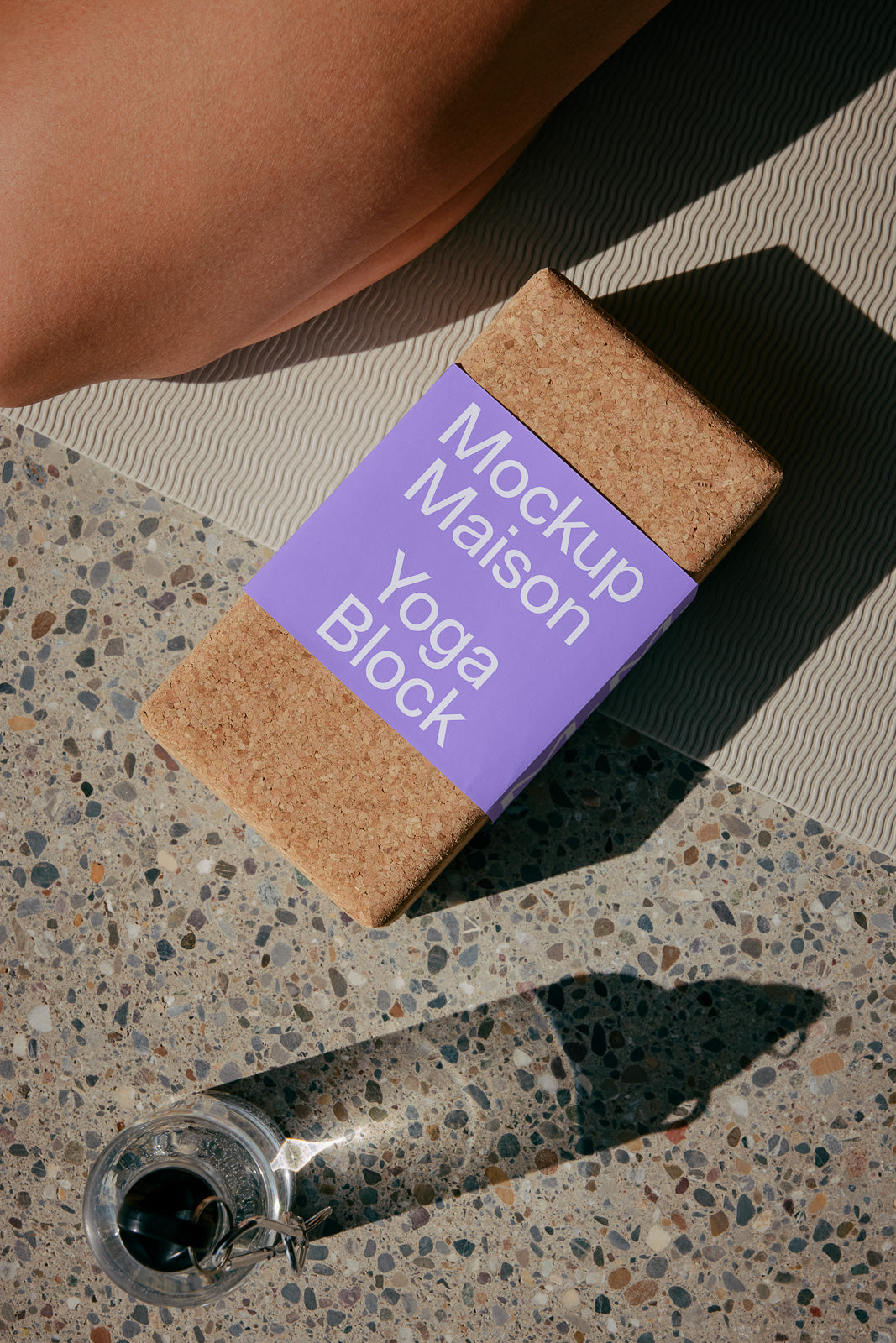 PSD mockup of a cork yoga block wrapped with a purple label that reads "Mockup Maison Yoga Block," placed on a textured surface near a yoga mat and glass of water.